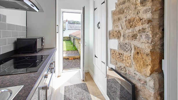 A modern kitchen with a stone wall, a view of the garden, and a door leading outside. - Host & Stay | Rose Cottage (Thropton)