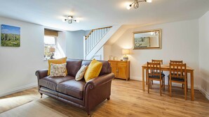 A living room with a brown leather sofa, wooden dining table, and chairs. There is a staircase with wooden railings and a window with a view of greenery. - Host & Stay | Rose Cottage (Thropton)