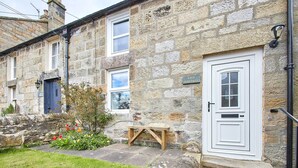A stone cottage with a white door, a small garden with red flowers, and a wooden bench. - Host & Stay | Rose Cottage (Thropton)