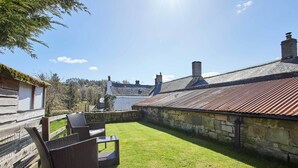 A courtyard with a grassy lawn, wicker furniture, stone walls, and a metal roof. - Host & Stay | Rose Cottage (Thropton)