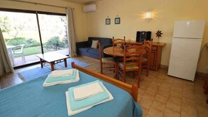 A room with a bed, a dining table with chairs, a sofa, a refrigerator, and a sliding glass door leading to a garden.
