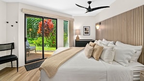 A modern bedroom with a large bed, a ceiling fan, a view of a garden through a sliding door, and a small desk with a chair. - Kauai Nihi Kai Villas 507 2 Bedroom Condo (Koloa)