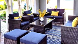An outdoor seating area with wicker furniture, cushions, and a central table with a vase of flowers.