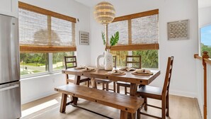 A modern kitchen with a wooden table, bench, and chairs, a refrigerator, and large windows with blinds.