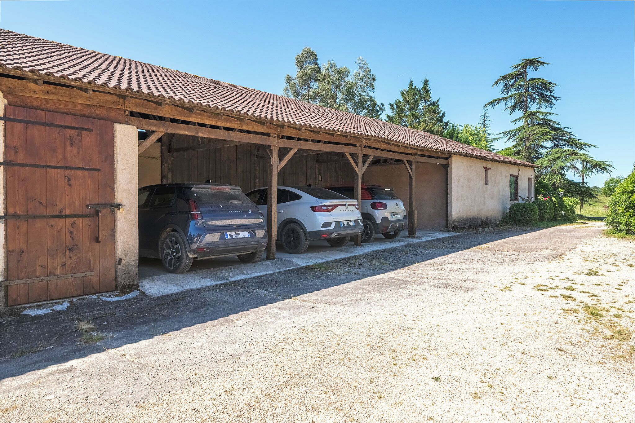 A covered carport with three cars parked inside, a gravel driveway, and a wooden building with a tiled roof.