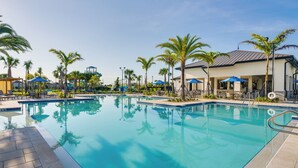 Piscina com espreguiçadeiras e guarda-sóis, rodeada por palmeiras e um edifício com varanda coberta.