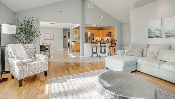 A spacious living room with a light-colored sectional sofa, a patterned armchair, and a round ottoman. A dining area with a table and chairs is visible in the background. - Benton Harbor Home w/ Fire Pit - 2 Mi to Beach! (Benton Harbor)