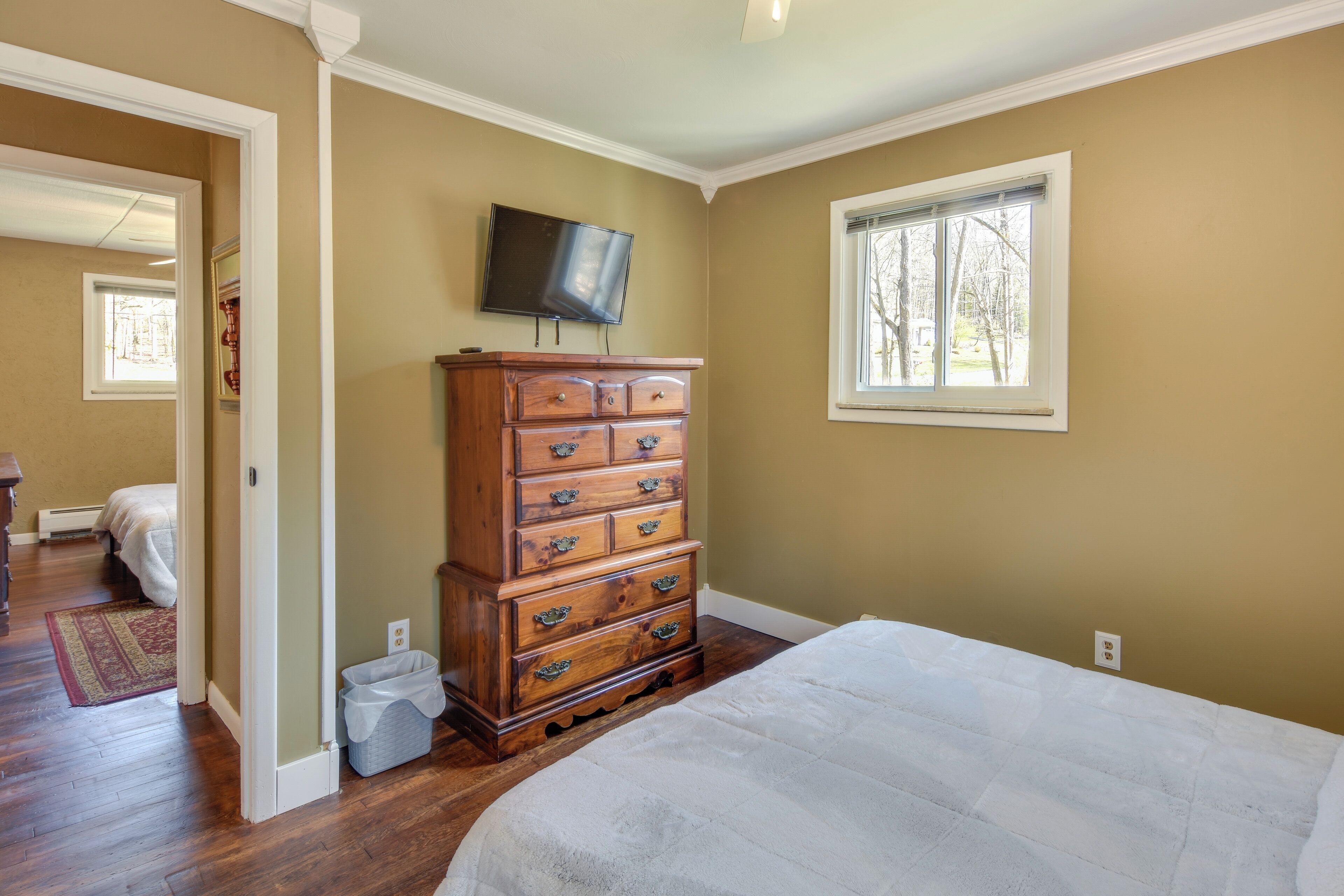 A bedroom with a large bed, a wooden dresser, a television mounted on the wall, and a window providing natural light.