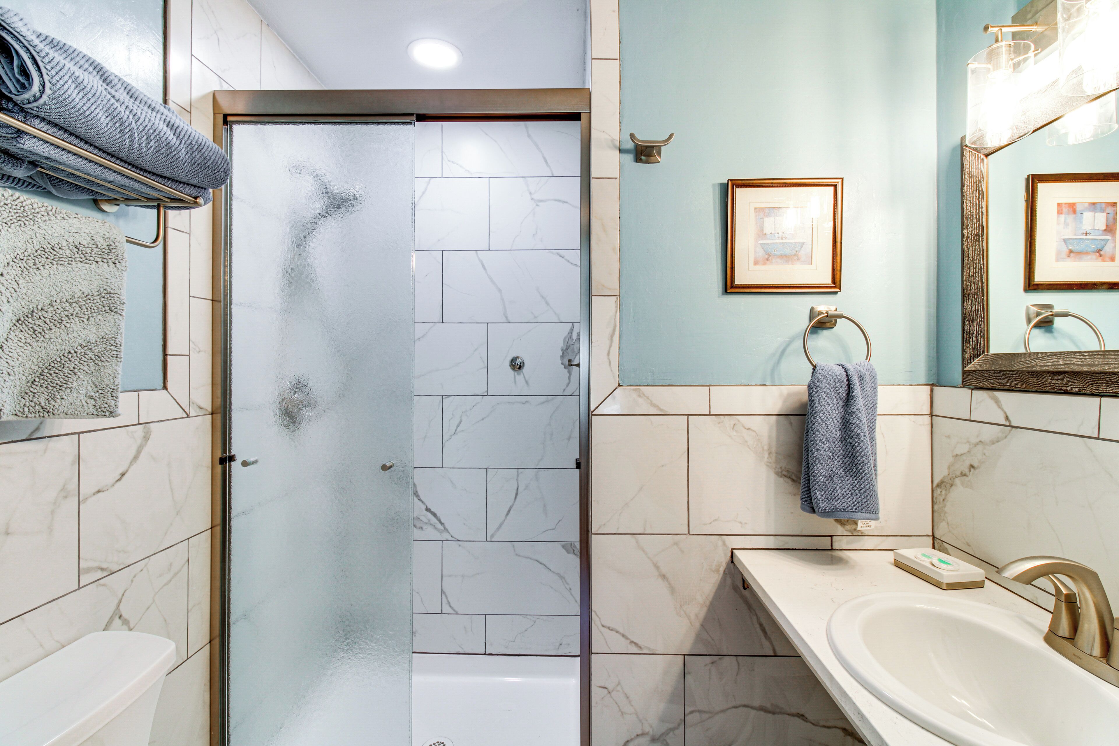 Une salle de bain avec une porte de douche en verre, un lavabo avec un robinet doré et deux tableaux encadrés au mur.