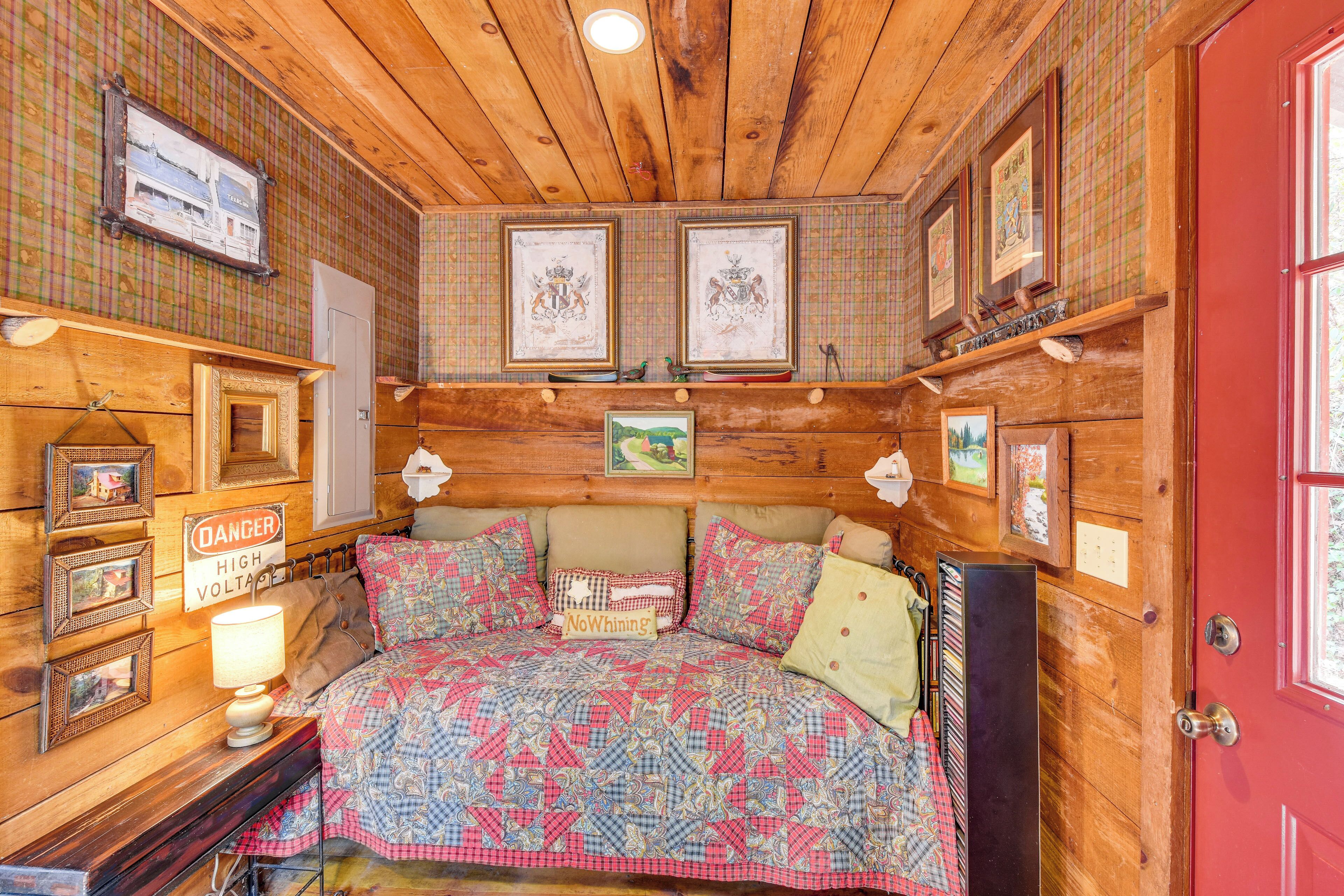 Un dormitorio tipo cabaña de madera con cama, cuadros enmarcados, un letrero de 'PELIGRO ALTA TENSIÓN' y un letrero que dice 'No Quejas'.