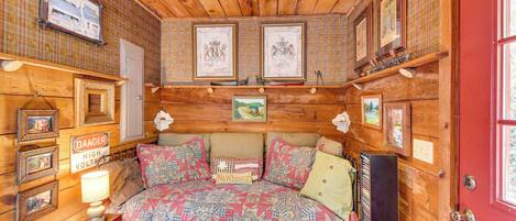Une chambre dans une cabane en bois, avec un lit, des photos encadrées, un panneau indiquant « DANGER : HAUTE TENSION » et un autre panneau indiquant « PAS DE PLAINTES ».