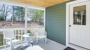 Een overdekte veranda met witte rieten stoelen en een kleine tafel, een groene deur, en uitzicht op bomen en een tuin.