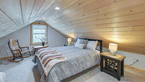 Een slaapkamer met een schuine kap, een bed met een beddegoed met dessin, een schommelstoel, een kleine tafel met een lamp, en een raam met jaloezieën.