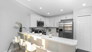 Una cocina moderna con gabinetes blancos, electrodomésticos de acero inoxidable y una zona de bar con taburetes blancos.