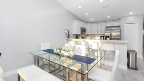 Una cocina moderna con armarios blancos, electrodomésticos de acero inoxidable y una mesa de comedor de vidrio con sillas blancas.