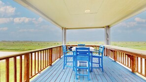 A covered outdoor deck with blue chairs and a table, overlooking a grassy landscape. - Seagull Shores: 1  BR, 1  BA House in Gilchrist, Sleeps 6 (Bolivar Peninsula)
