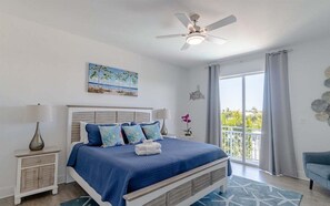 A bedroom with a bed, a nightstand, a ceiling fan, a window with curtains, and a view of greenery outside.