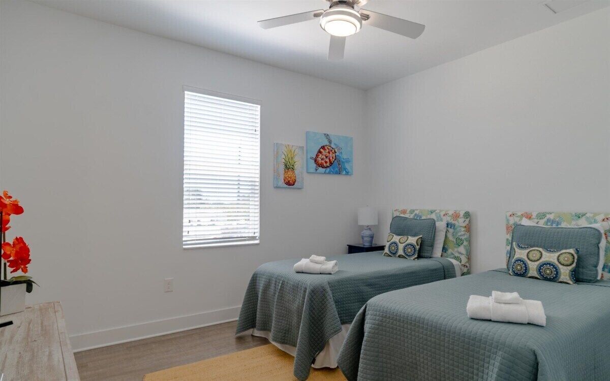 Un dormitorio con dos camas, un ventilador de techo, una ventana con persianas y dos cuadros decorativos.
