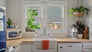 Una cocina con salpicadero blanco de azulejos, un lavamanos doble, microondas, una planta colgante y una ventana con vistas a zonas verdes.