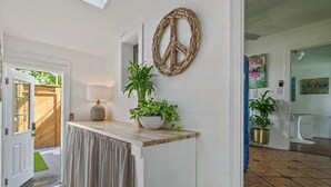 Un pasillo con pared blanca, puerta de madera, cortina color beige, banca de madera, planta en maceta, decoración con un símbolo de la paz y suelo de baldosas.