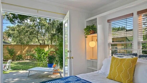 Un dormitorio con cama, una ventana con persianas, una mesita de noche y vistas a un patio trasero con terraza y plantas.