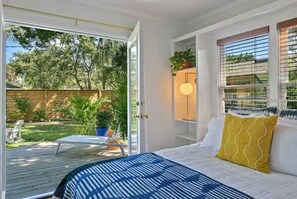 Een slaapkamer met een bed, een raam met zonwering, een nachtkastje en uitzicht op een achtertuin met een terras en planten.