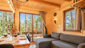 Interno di una baita di legno con un tavolo da pranzo per quattro persone, un divano grigio e ampie finestre con vista sulla foresta.
