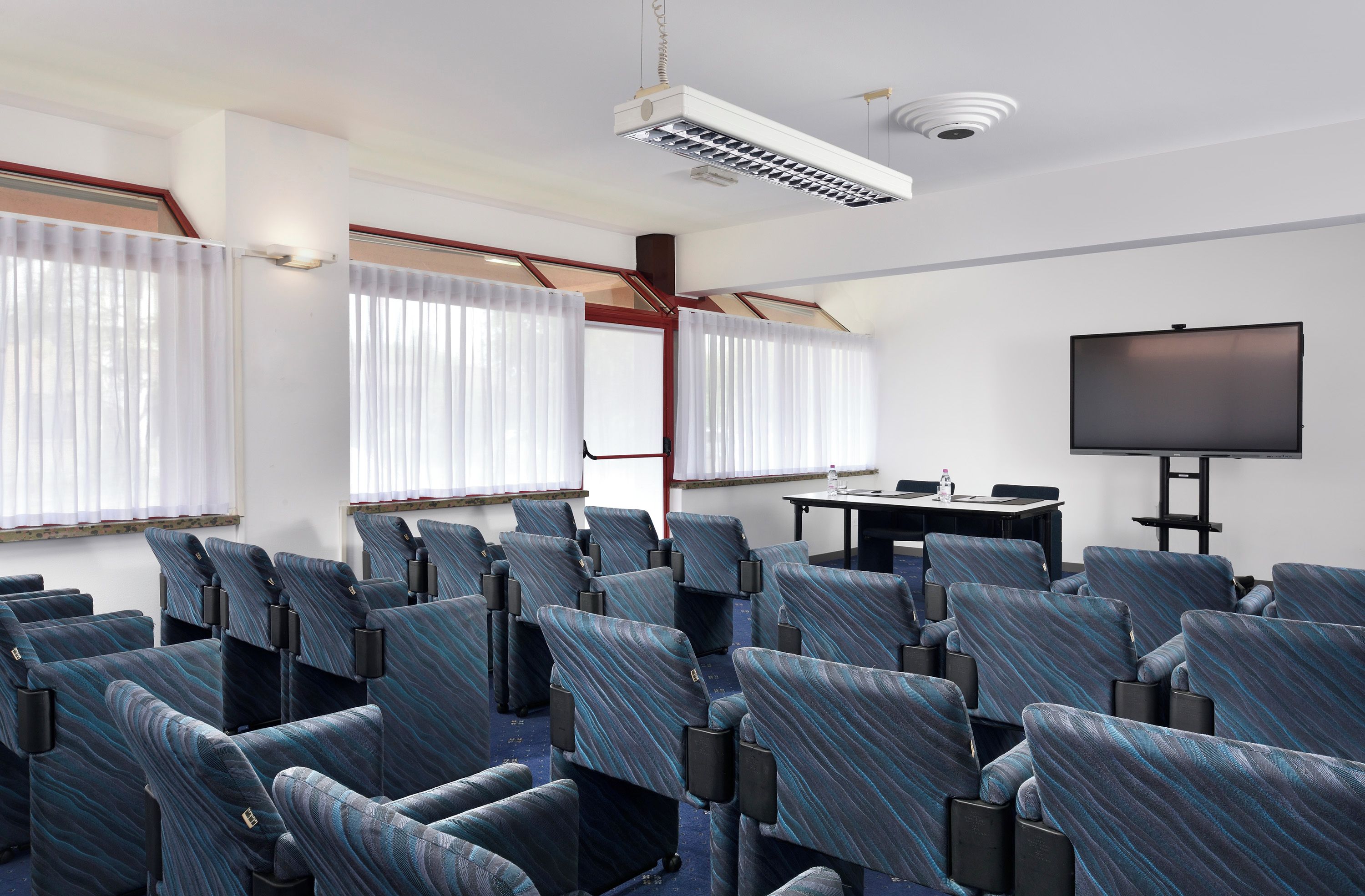 Ein Konferenzraum mit Reihen von Stühlen, einem Projektor, einer Leinwand und einem Tisch mit einer Wasserflasche.