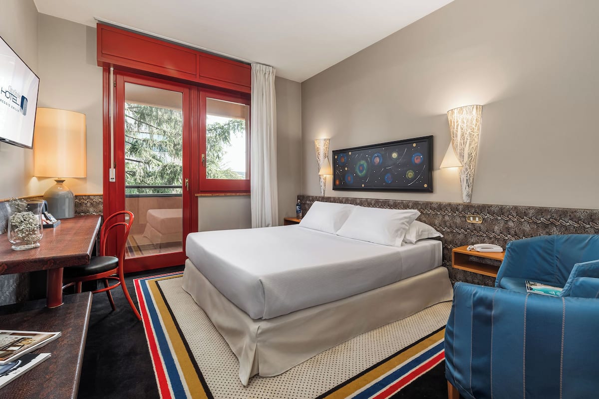 Ein Hotelzimmer mit einem Bett, einem Schreibtisch mit Stuhl, einem blauen Sessel, einem Fernseher und einem Fenster mit roter Tür.