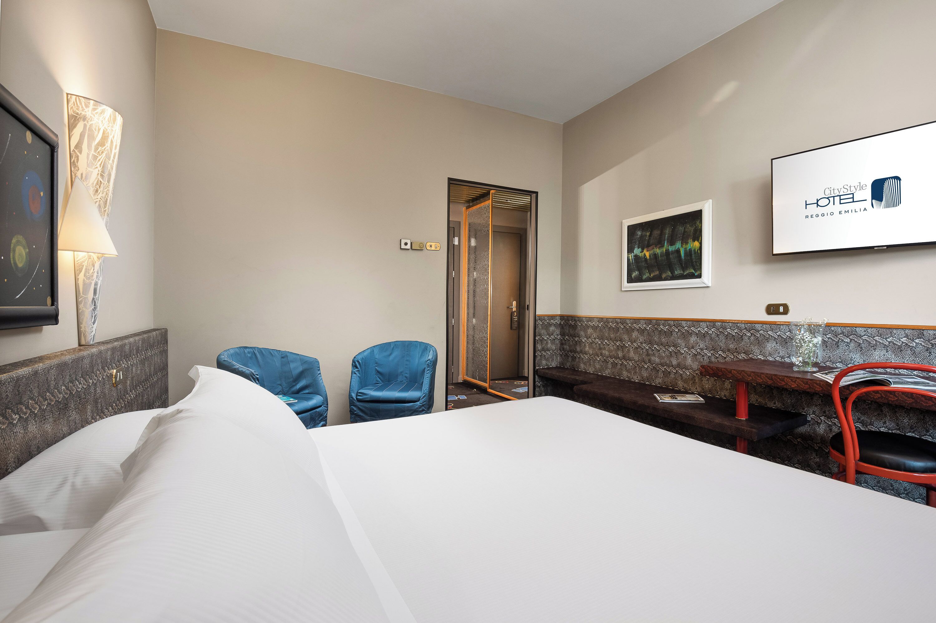 A hotel room with a bed, two blue chairs, a desk with a red chair, a TV on the wall, and a framed picture.
