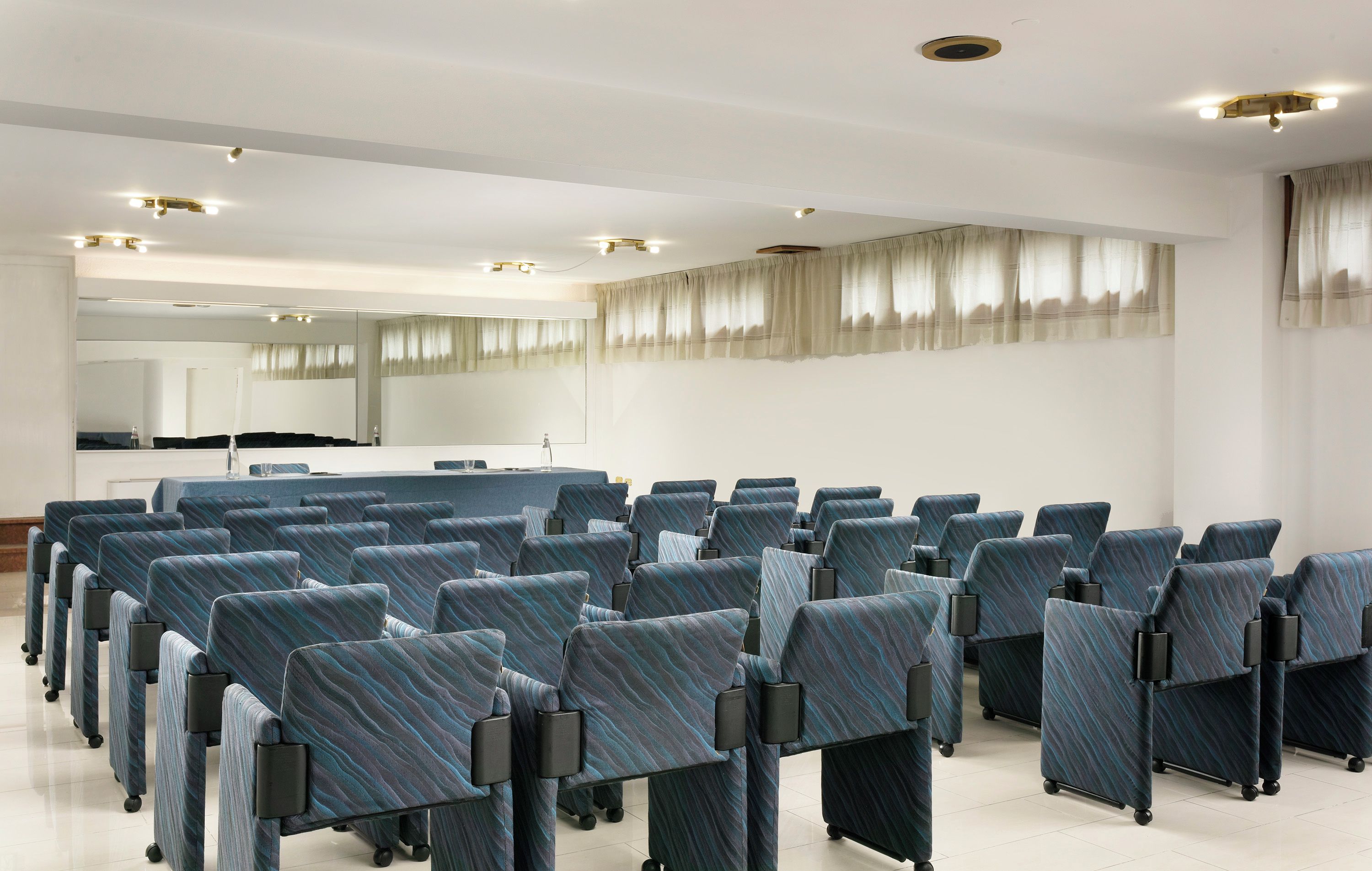 Un salón de conferencias con filas de sillas, una mesa grande y un espejo en la pared.