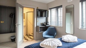 Una habitación de hotel moderna con una cama grande, un área de descanso con una silla, una kitchenette pequeña y un baño visible a través de una puerta abierta.