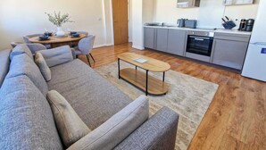A modern kitchen with stainless steel appliances, a dining area with a round table and grey upholstered chairs, and a living room with a grey sofa and a wooden coffee table. - 2 Bed City Centre Flat | Free Parking (Manchester)