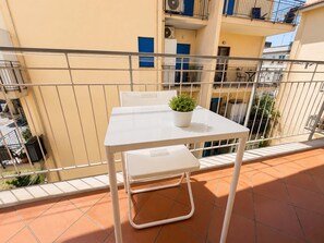 Un balcón con una mesa y una silla blancos, una planta en maceta y vistas de edificios vecinos.