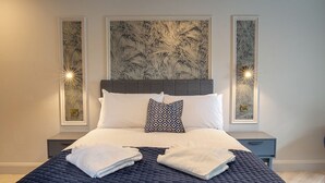 Un lit bien fait, recouvert d’une couverture sombre, avec des oreillers blancs et un plaid à motifs. On y trouve deux tables de chevet avec des lampes, et des œuvres d’art ornent les murs.