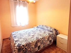 Une petite chambre à coucher avec un lit, une table de chevet et une fenêtre ornée d’un rideau léger.