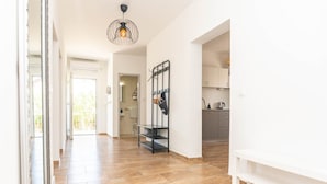 A hallway with a wooden floor, a hanging light fixture, and a kitchen area in the background.