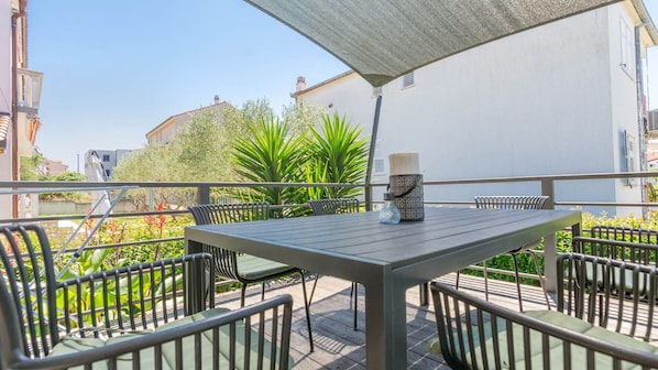 An outdoor patio with a table and chairs, surrounded by greenery and a white building in the background.