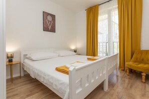 A white bed with a headboard, a bedside lamp, a chair, and a window with yellow curtains.
