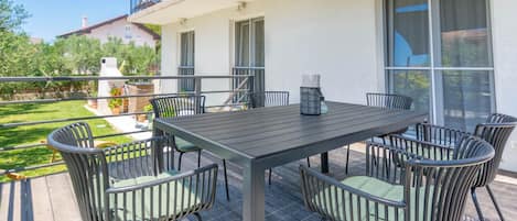 Een terras met een tafel, stoelen, een hekwerk en uitzicht op een tuin en gebouwen.