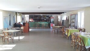 A spacious room with multiple tables and chairs, a bar area with shelves of bottles, and a view of the outside.