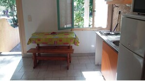 A small kitchen with a wooden table and bench, a refrigerator, and a window with a view of greenery.