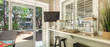 A covered patio area with a wicker chair, a small stool, and a table with a lantern and a bowl.