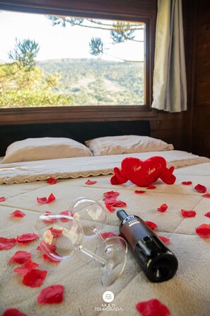 A bed with a white bedspread, a red heart-shaped pillow, and scattered rose petals. - Chalé Diamante Negro - Urubici - SC (Urubici)