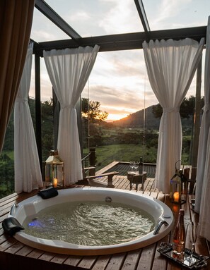 Un bain à remous offrant une vue sur un coucher de soleil se déclinant sur un paysage, agrémenté de rideaux et de bougies.