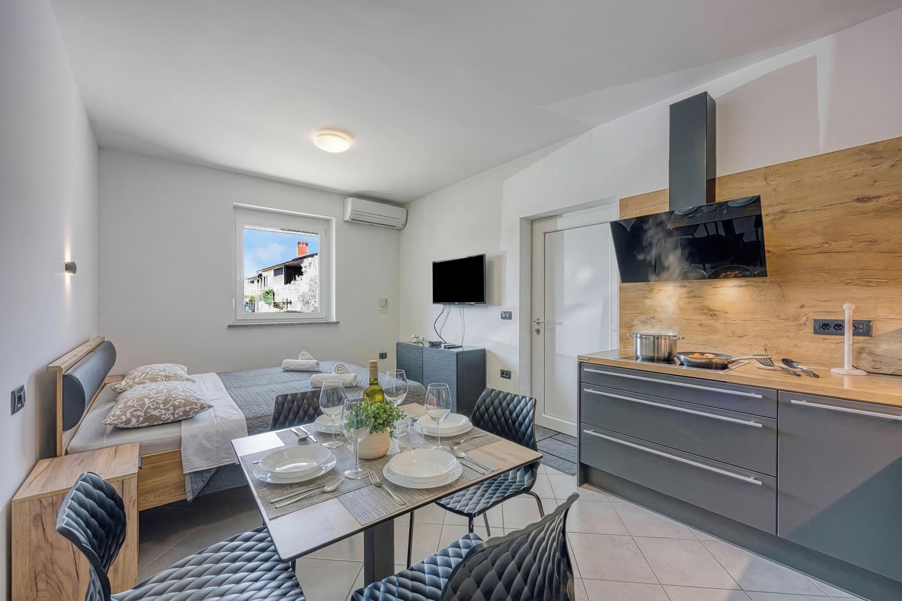 Habitación de hotel compacta con cama, mesa de comedor para dos personas, kitchenette, televisor montado en la pared y ventana con vista a un edificio.
