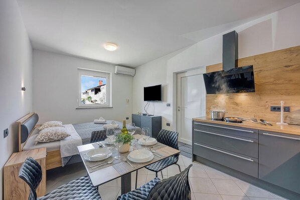 Quarto de hotel pequeno com cama, mesa de jantar para dois, cozinha compacta, televisão presa à parede e janela com vista para um prédio.