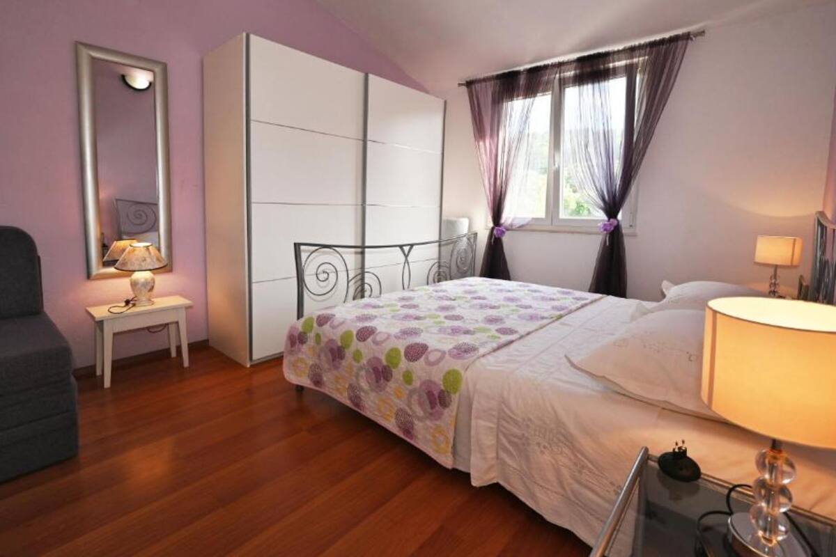 A bedroom with a bed, a chair, a wardrobe, a bedside table, and a window with curtains.
