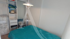 Et lille, pænt møbleret værelse med en seng dækket af et teal farvet tæppe, en hvid garderobe og et skrivebord med en lampe.