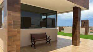 An outdoor seating area with a bench and a large window overlooking a green landscape. - Walk to Palm Beach • Pool • Modern 2BR Aruba Condo (Noord)